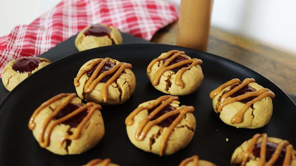 A black plate holds several peanut butter cookies filled with jelly and drizzled with peanut butter. More cookies and a red checkered cloth are in the background on a wooden surface.