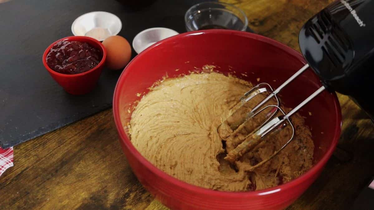Beaters mixing light brown batter in a red bowl sitting on a table with a slate board that has condiment dishes of other ingredients on it visible in the background.