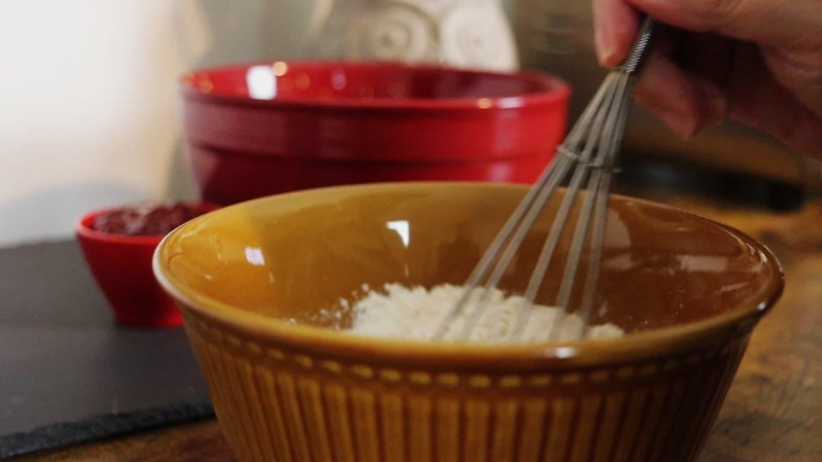 Whisk in a brown bowl with some flour visible at the top of the bowl and a red bowl in the background.