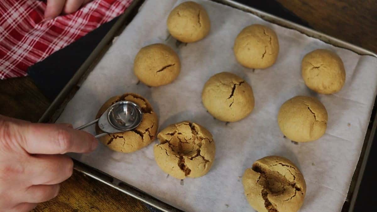 Creating indents in the top of peanut butter cookie balls sitting on a parchment lined baking sheet with a metal measuring spoon.