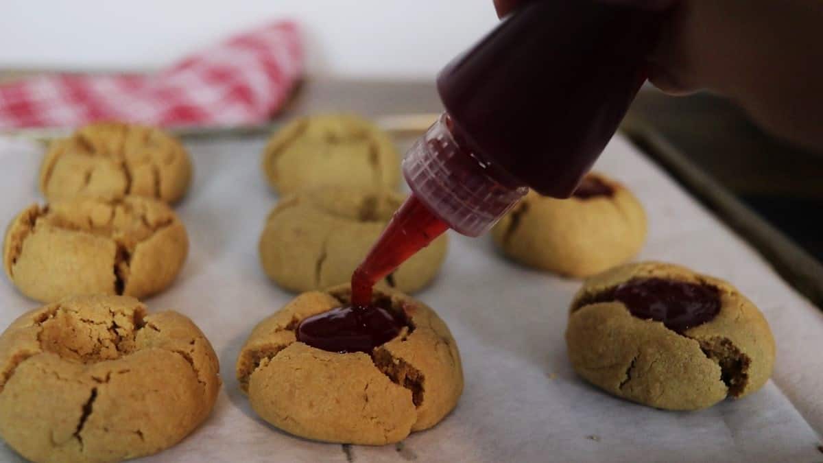 Jelly being piped from a squeeze bottle onto the top of a peanut butter cookie sitting on parchment paper