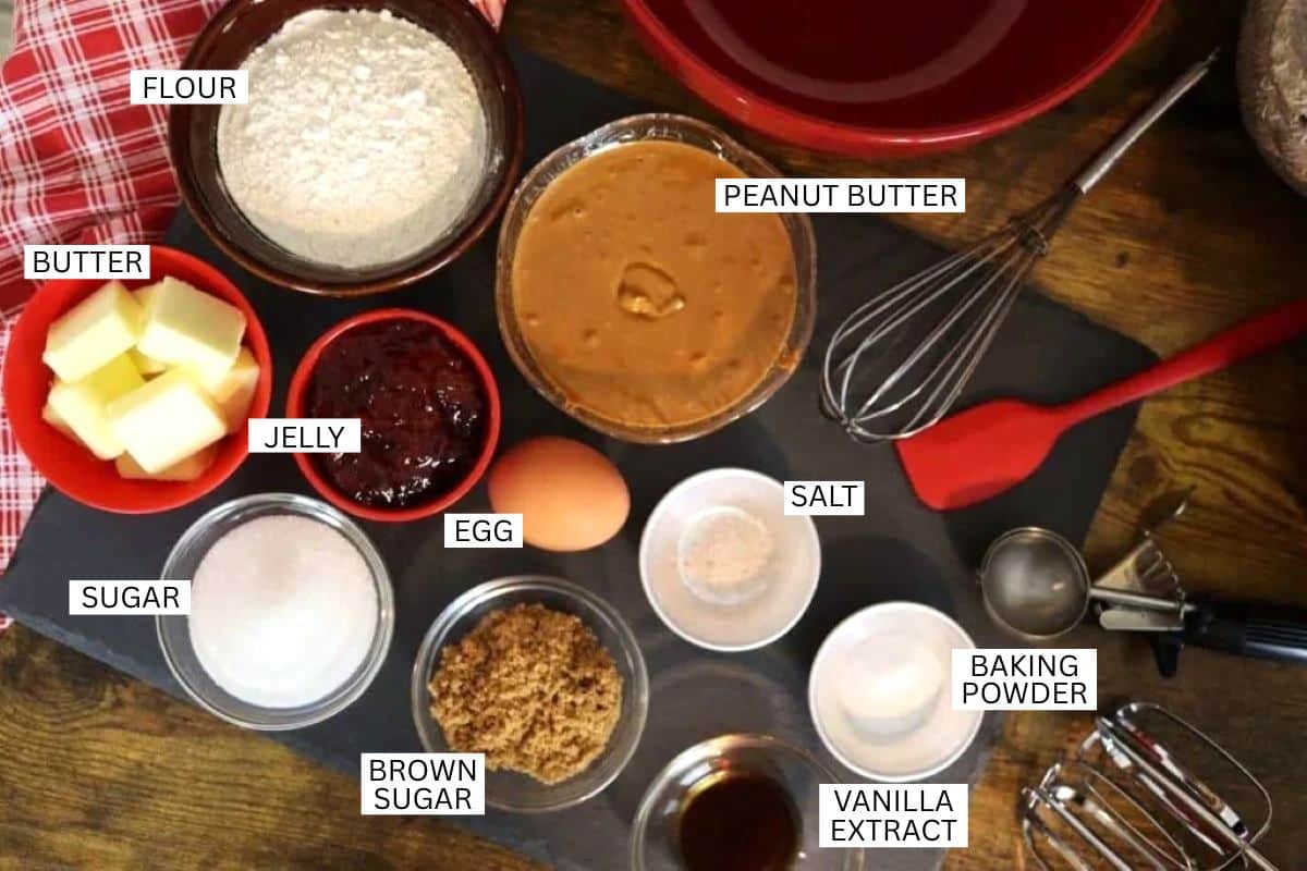 Baking ingredients labeled in bowls and dishes, including flour, peanut butter, butter, jelly, egg, sugar, brown sugar, salt, baking powder, and vanilla extract, arranged on a kitchen counter.