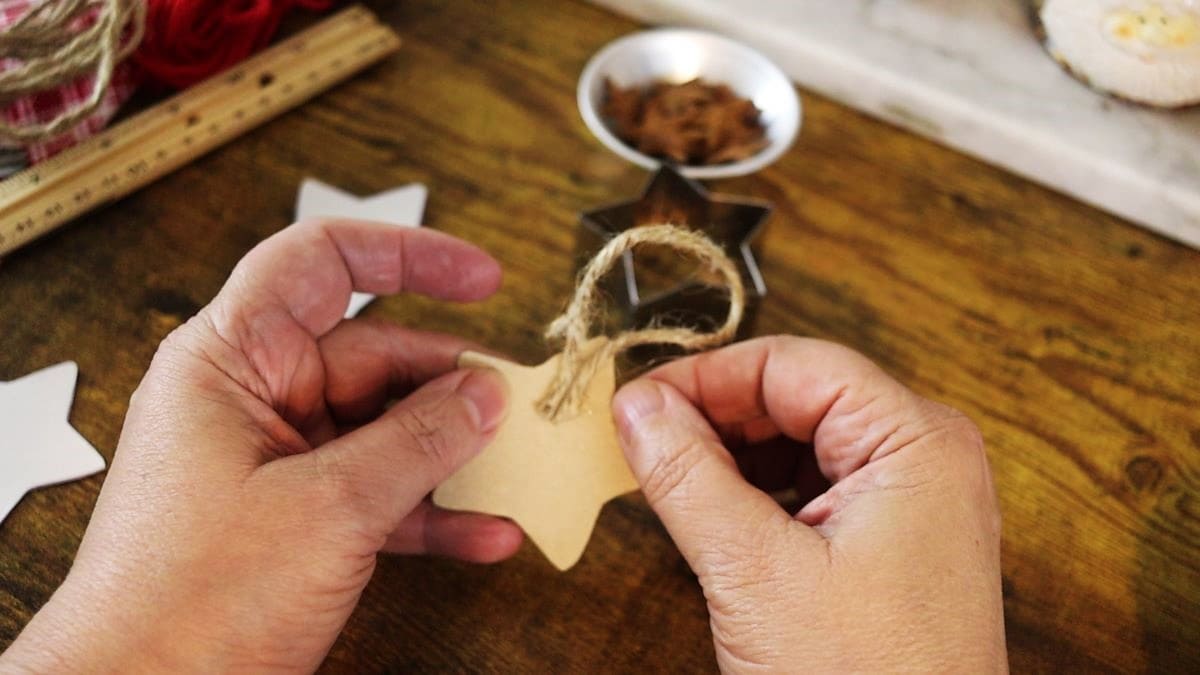 Hands holding a cardboard star cutout with a jute twine loop attached to the back of it. There are other craft supplies visible in the background.