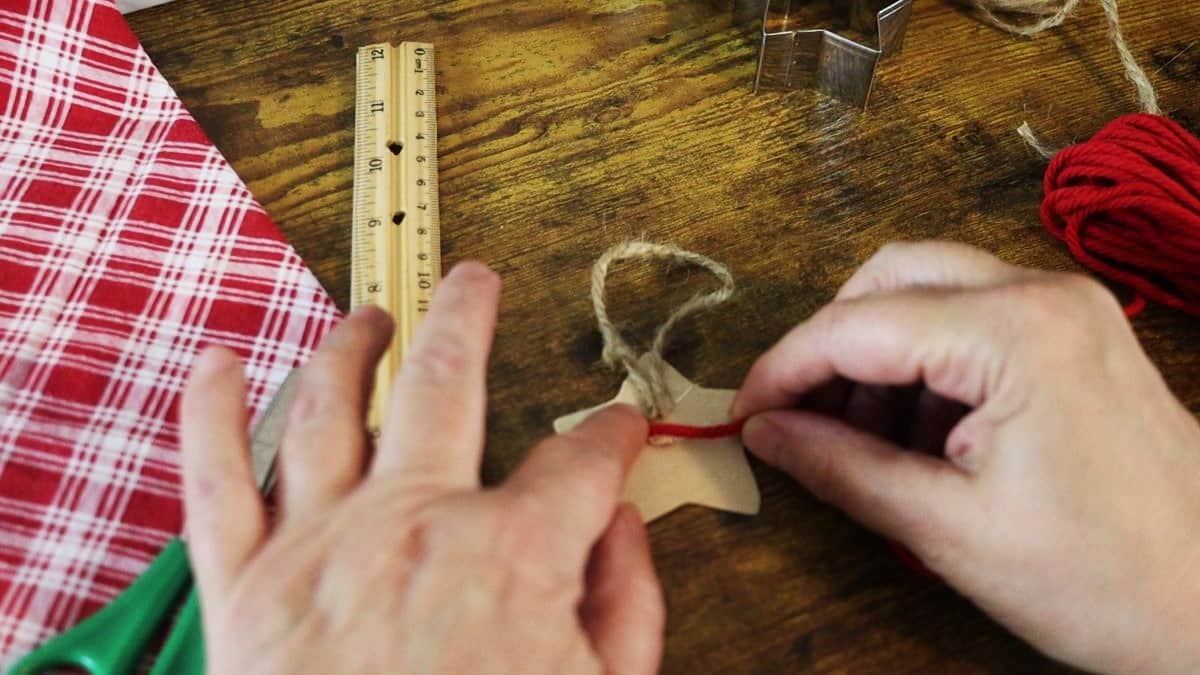 Hands attaching a piece of red cord to the back of a cardboard star cutout. There is a ruler, a red checkered cloth, and a cookie cutter visible to the sides of it.