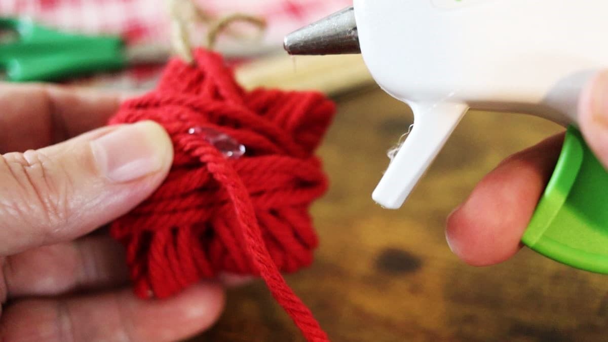 The tip of a hot glue gun above a red twine star ornament with hot glue visible on the twine.