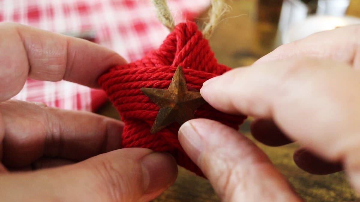 Hands placing a rustic metal star in the center of a star ornament made from red cord.