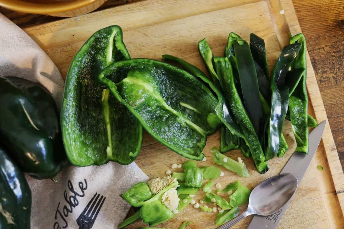 Halved and sliced poblano peppers on a wooden cutting board with a spoon, knife, seeds, and stem pieces.