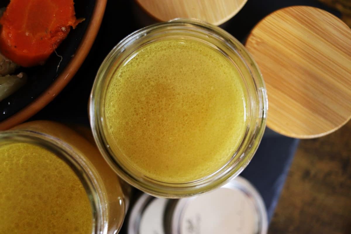A glass jar filled with yellow liquid, possibly broth, surrounded by wooden lids and part of a bowl containing vegetables like a carrot.