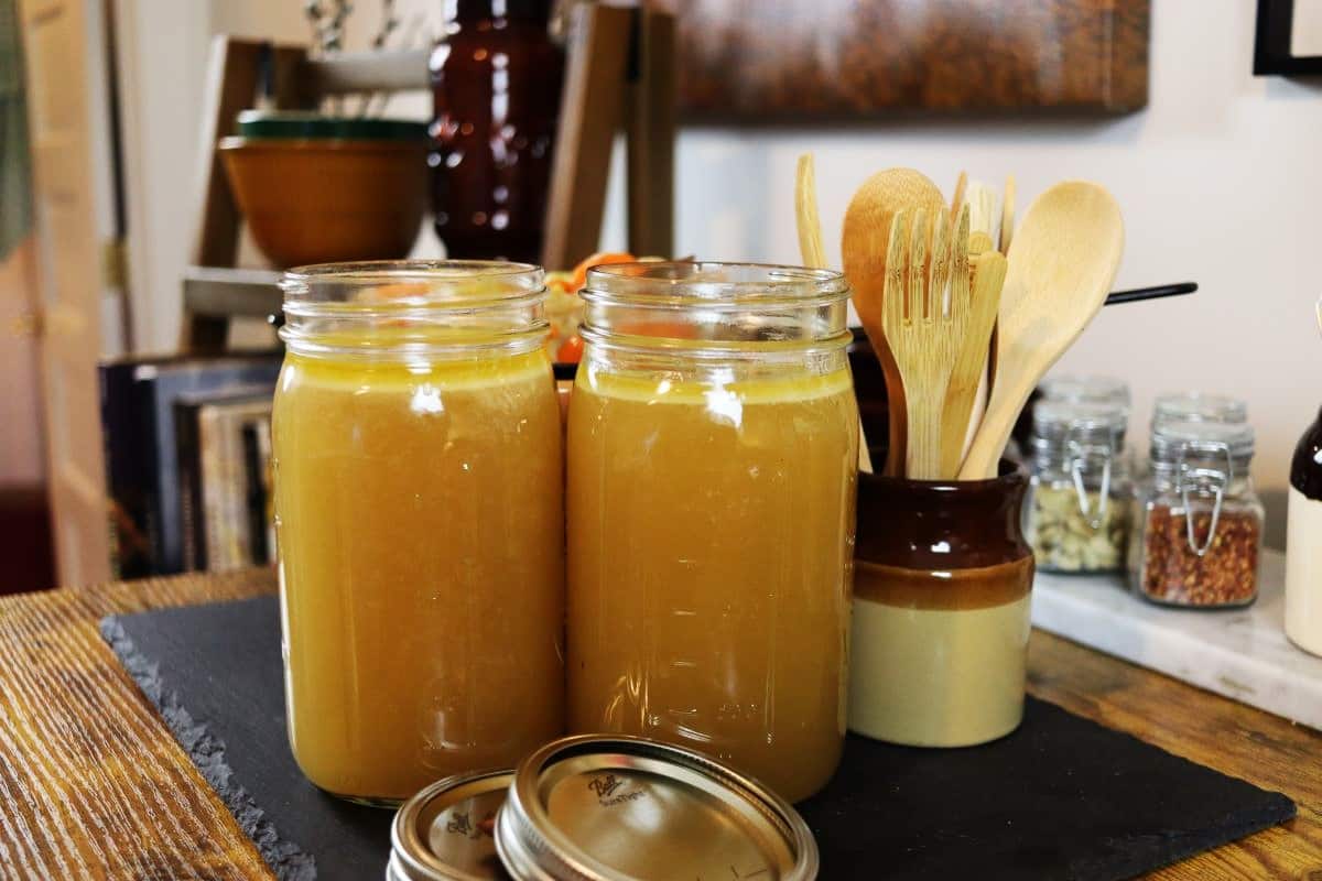 two mason jars of homemade chicken broth on a counter with lids sitting in front of them and a jar of wooden utensils sitting behind them.