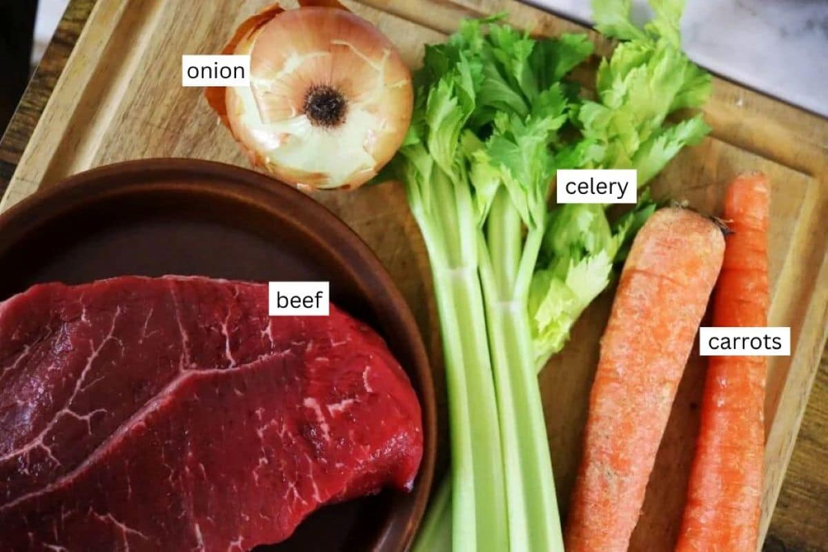 A wooden cutting board with raw beef, a halved onion, celery stalks, and two carrots, each labeled with text indicating beef, onion, celery, and carrots.