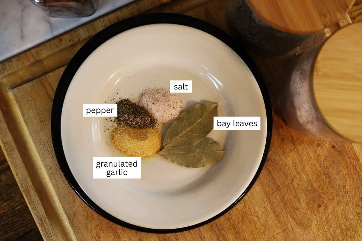 A white plate with a black rim holds grouped seasonings: ground pepper, granulated garlic, salt, and two bay leaves. Each spice is labeled. The plate sits on a wooden surface next to jars.