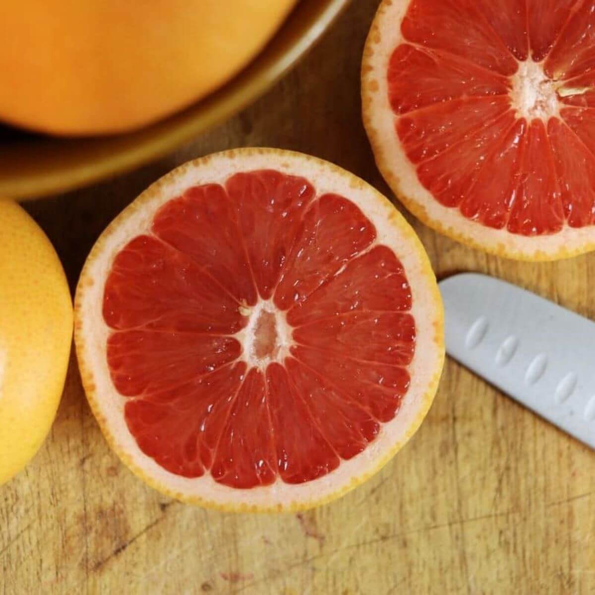 Halved pink grapefruit showing juicy red flesh on a wooden cutting board beside a knife.
