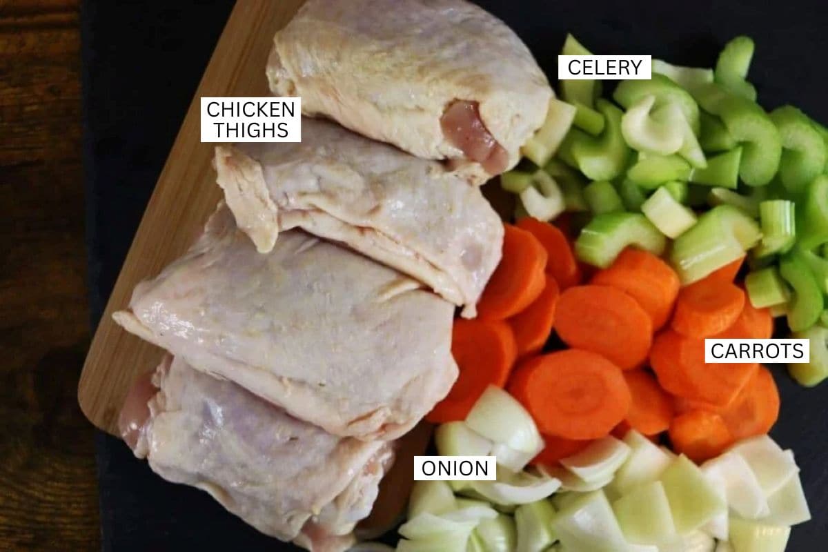 Raw chicken thighs, sliced carrots, chopped celery, and diced onions are laid out on a cutting board, each ingredient labeled with text.