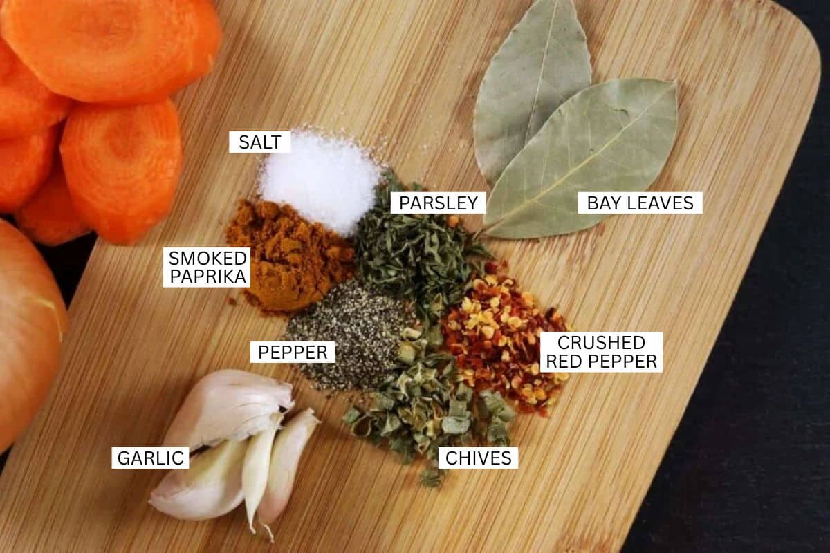 A wooden cutting board with labeled piles of spices and herbs: salt, parsley, bay leaves, smoked paprika, pepper, crushed red pepper, chives, and garlic. Sliced carrots and onion are partially visible on the left.