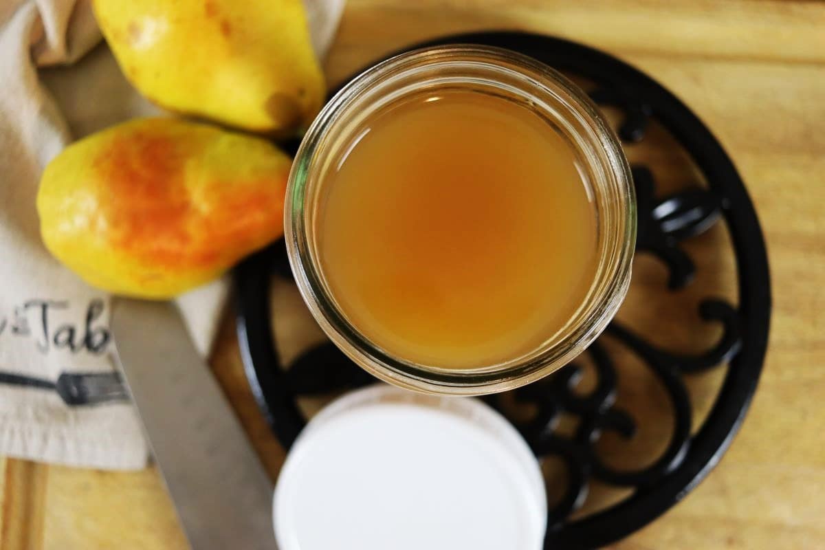 A top-down view of a jar filled with light brown liquid, next to two yellow pears, a knife, and a white lid on a wooden surface with a decorative black trivet underneath the jar.