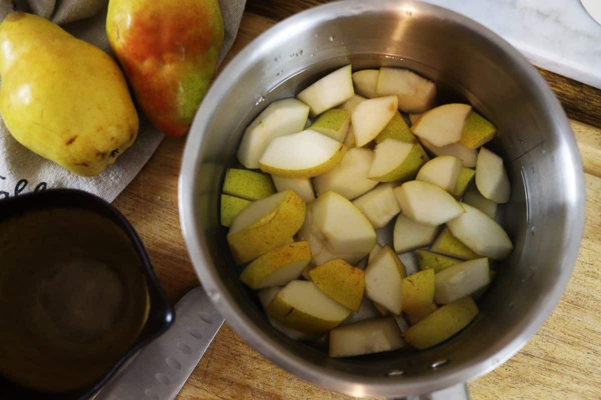 Pear chunks and water in a saucepan with whole pears visible to the side of it.