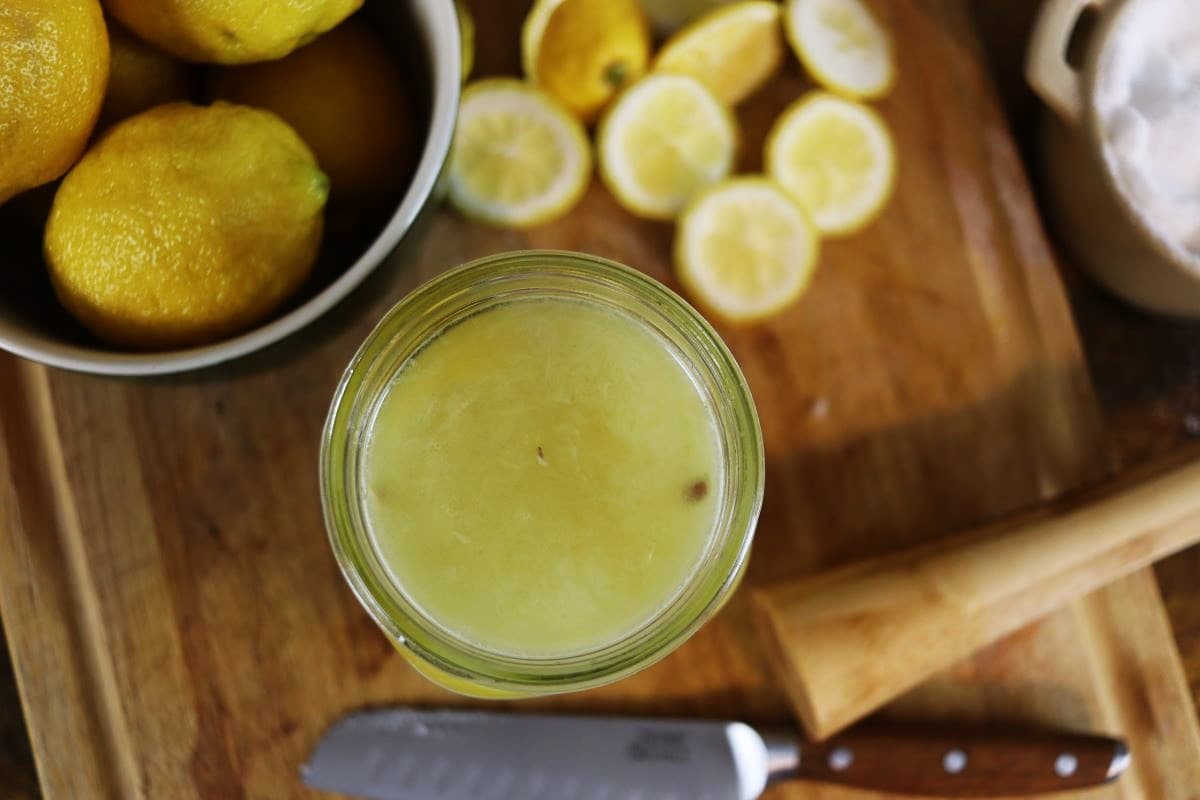 Jar of yellow colored liquid on a wooden surface with a knife, lemon ends and a bowl of lemons visible around it.