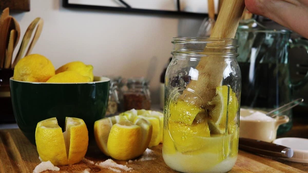 Wooden sauerkraut pounder pushing lemons into a mason jar with salt and liquid visible in the bottom. There are quartered lemons, a bowl of lemons, and kitchen decor visible in the backgroudn.