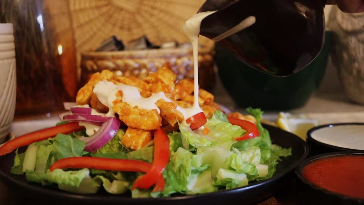drizzling homemade blanch dressing over a salad topped with red bell peppers, red onion, and chunks of chicken covered in buffalo sauce.