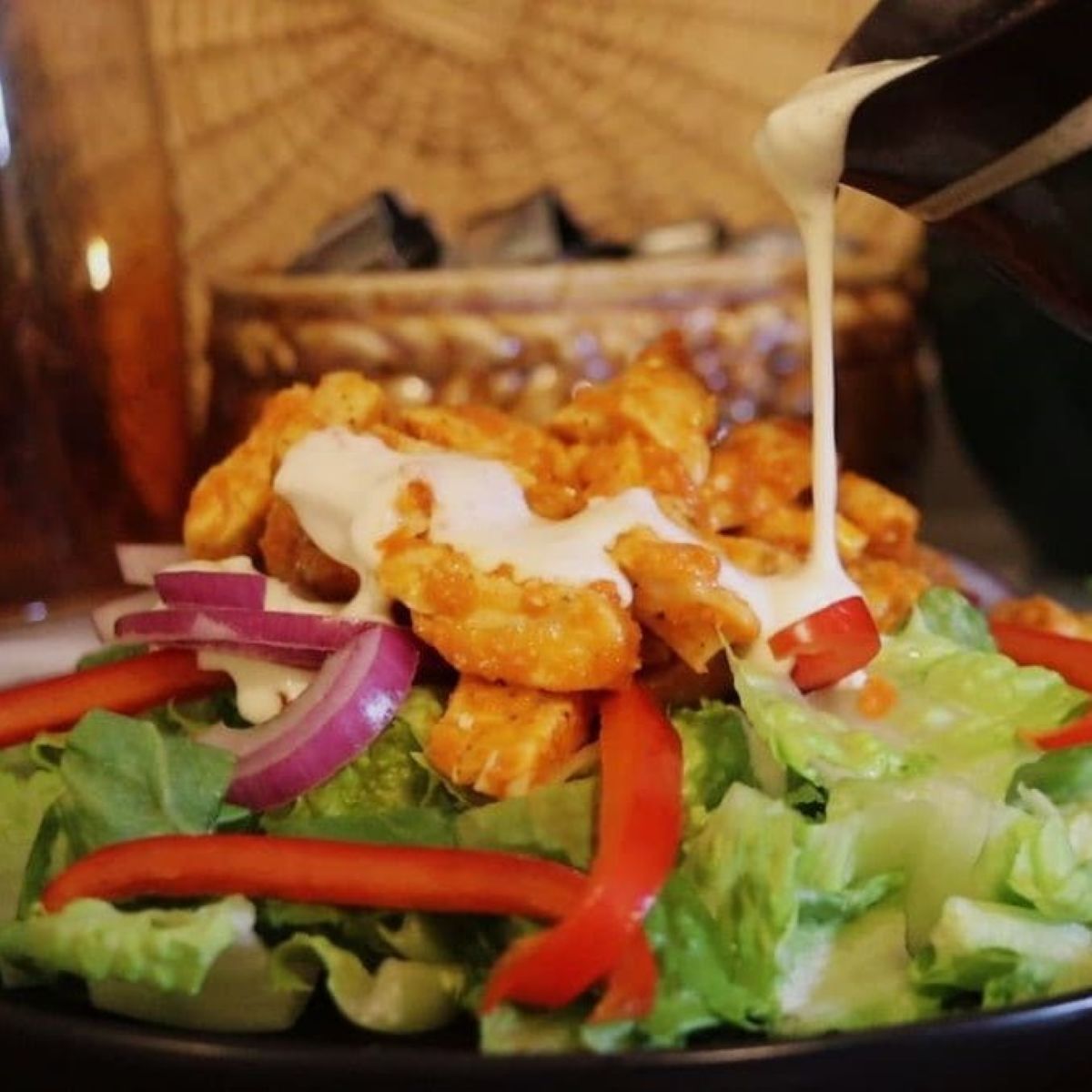 A salad with lettuce, red onion, and red bell pepper, topped with pieces of buffalo chicken, as creamy dressing is being poured over it.