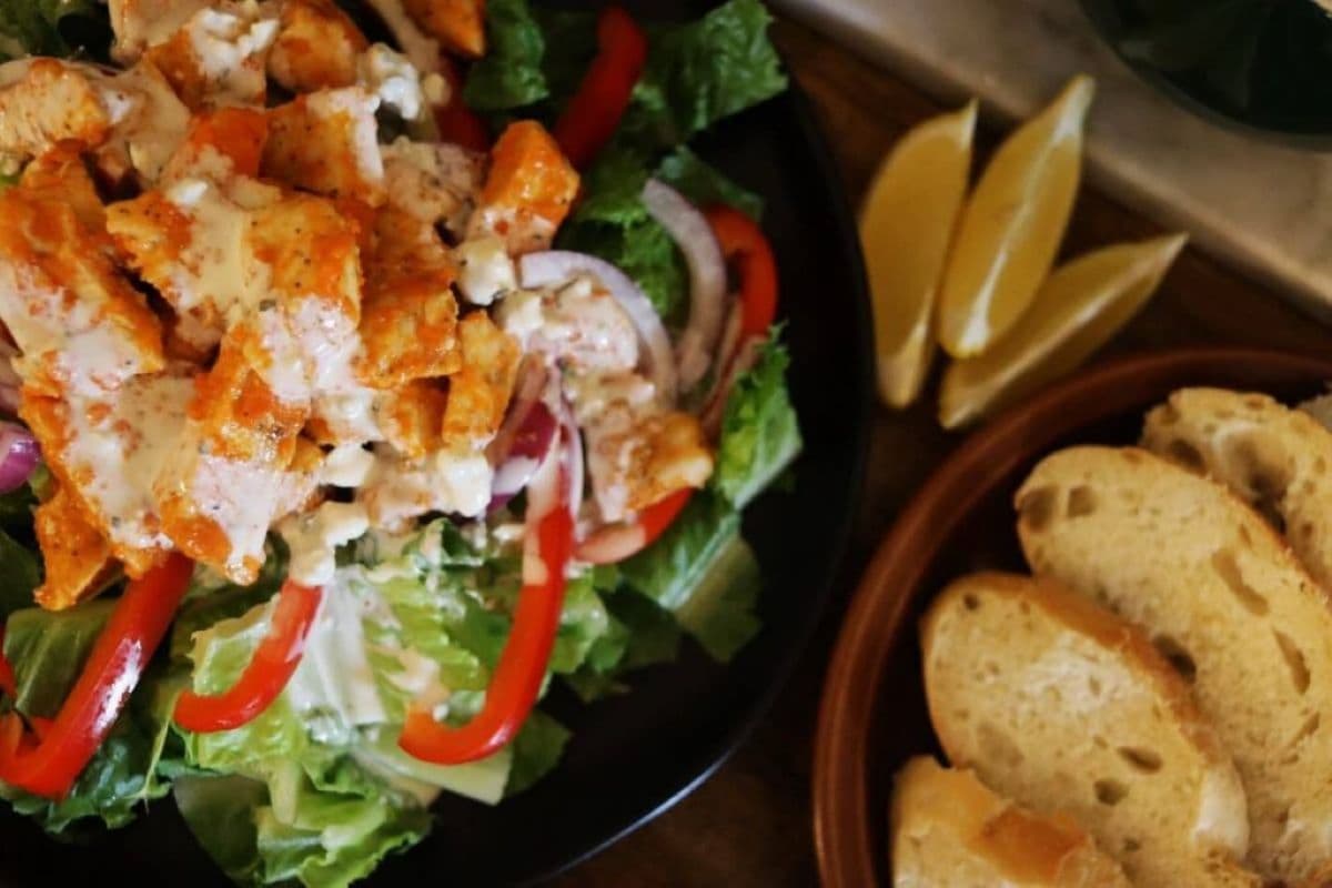 A plate of salad with lettuce, red bell peppers, onions, grilled buffalo chicken pieces, and creamy dressing, next to a dish of lemon wedges and a plate of sliced bread.