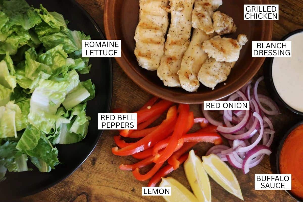 A tabletop with labeled ingredients: a bowl of romaine lettuce, strips of grilled chicken, sliced red bell peppers, sliced red onion, lemon wedges, buffalo sauce, and a bowl of ranch dressing.