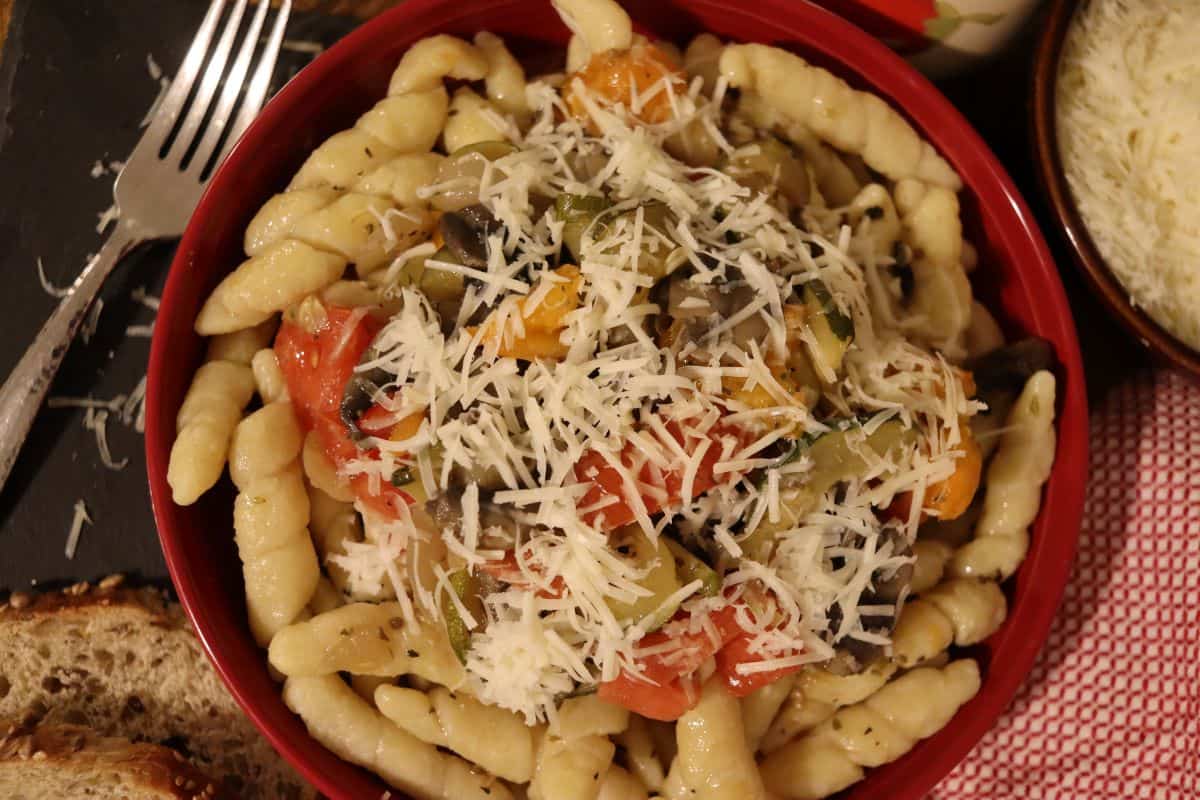 A bowl of pasta topped with sautéed vegetables and grated cheese, served with slices of bread on the side and a fork nearby.