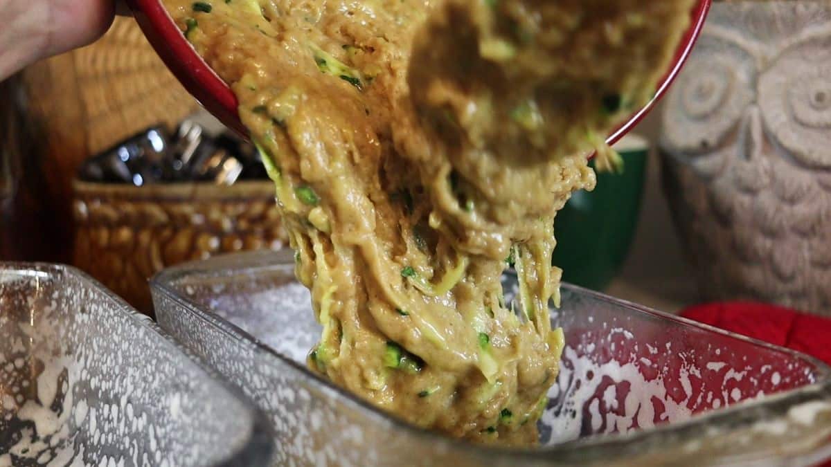 Batter being poured from a bowl into a greased loaf pan with kitchen utensils and decor visible in the background.