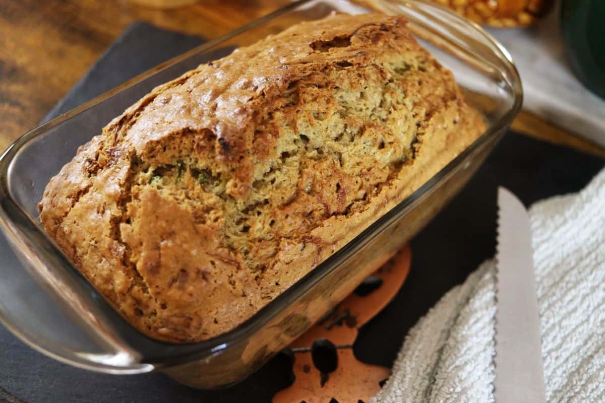 Golden brown loaf of bread in a loaf pan with a bread knife and a towel next to it.