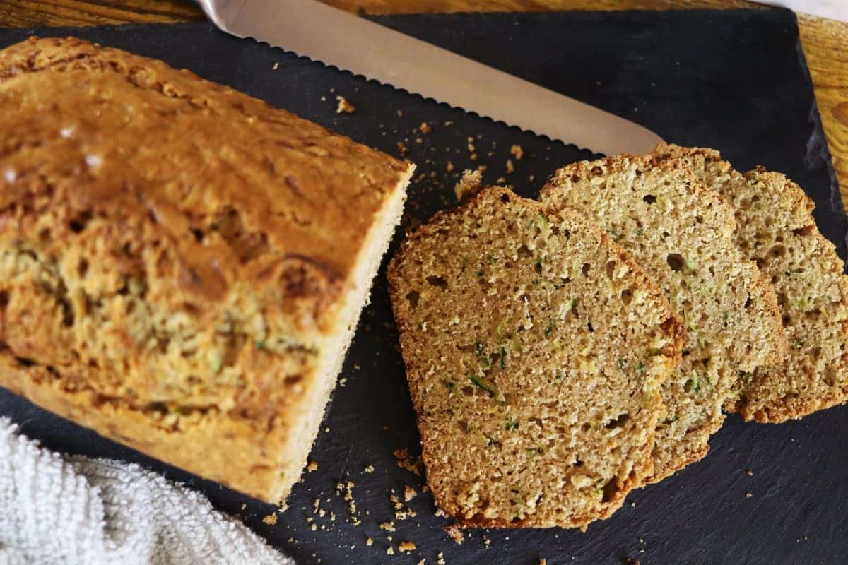 A loaf of zucchini bread on a dark cutting board, partially sliced with three pieces laid out next to the unsliced portion. A serrated knife and a textured cloth are beside the bread.