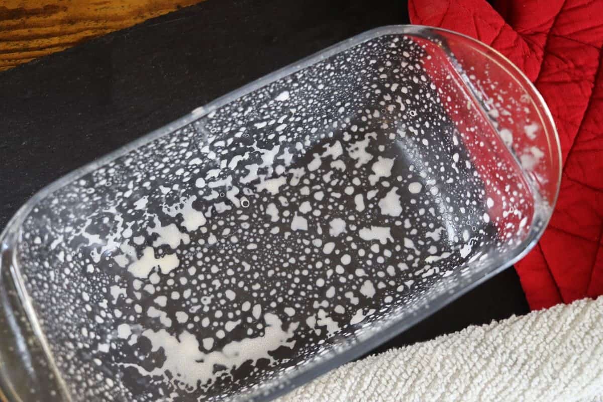 Greased glass loaf pan on a slate board with red oven mitts and a light green towel next to it.