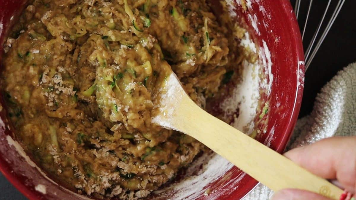 Batter with shredded zucchini visible in it in a red bowl with a wooden spoon in the bowl.