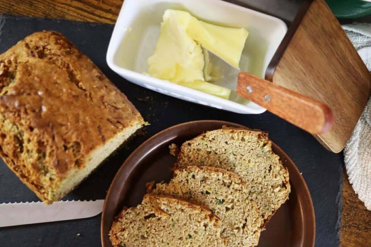 A loaf of bread with several slices cut sits on a brown plate next to a butter dish containing butter. A wooden-handled knife is nearby on a dark surface.