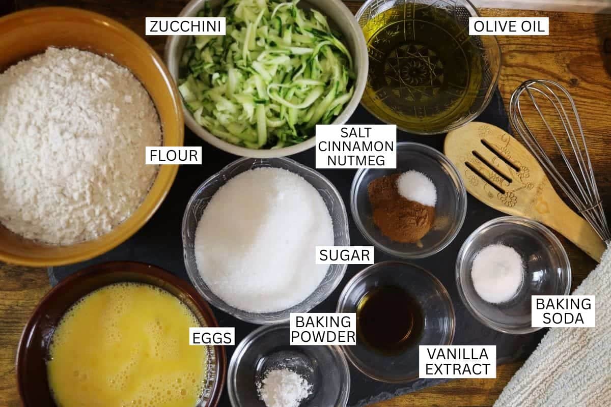 Labeled baking ingredients in bowls: flour, shredded zucchini, olive oil, sugar, eggs, baking powder, vanilla extract, baking soda, and a bowl with salt, cinnamon, and nutmeg. A whisk and wooden spatula are beside them.