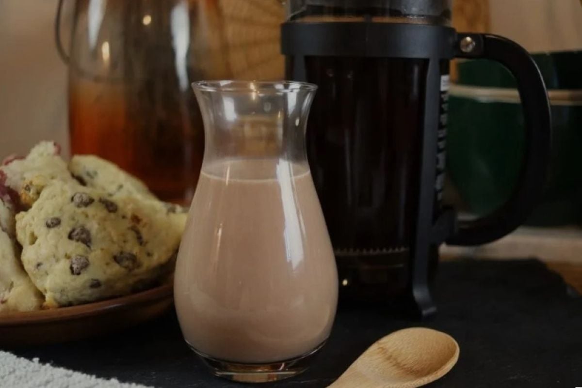 A small glass carafe filled with chocolate creamer sits on a table beside a French press, a wooden spoon, and a plate of chocolate chip scones. The background is softly lit and cozy.