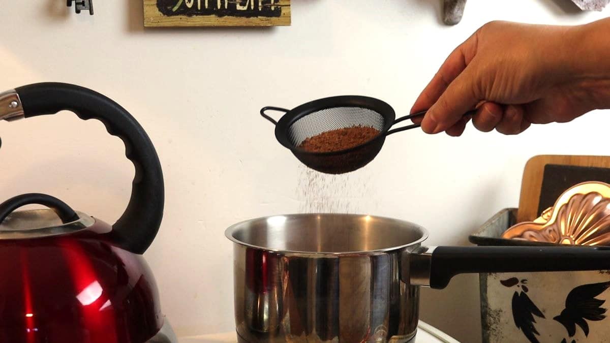 Small saucepan sitting on a stovetop with cocoa being sifted into it from a small mesh strainer.