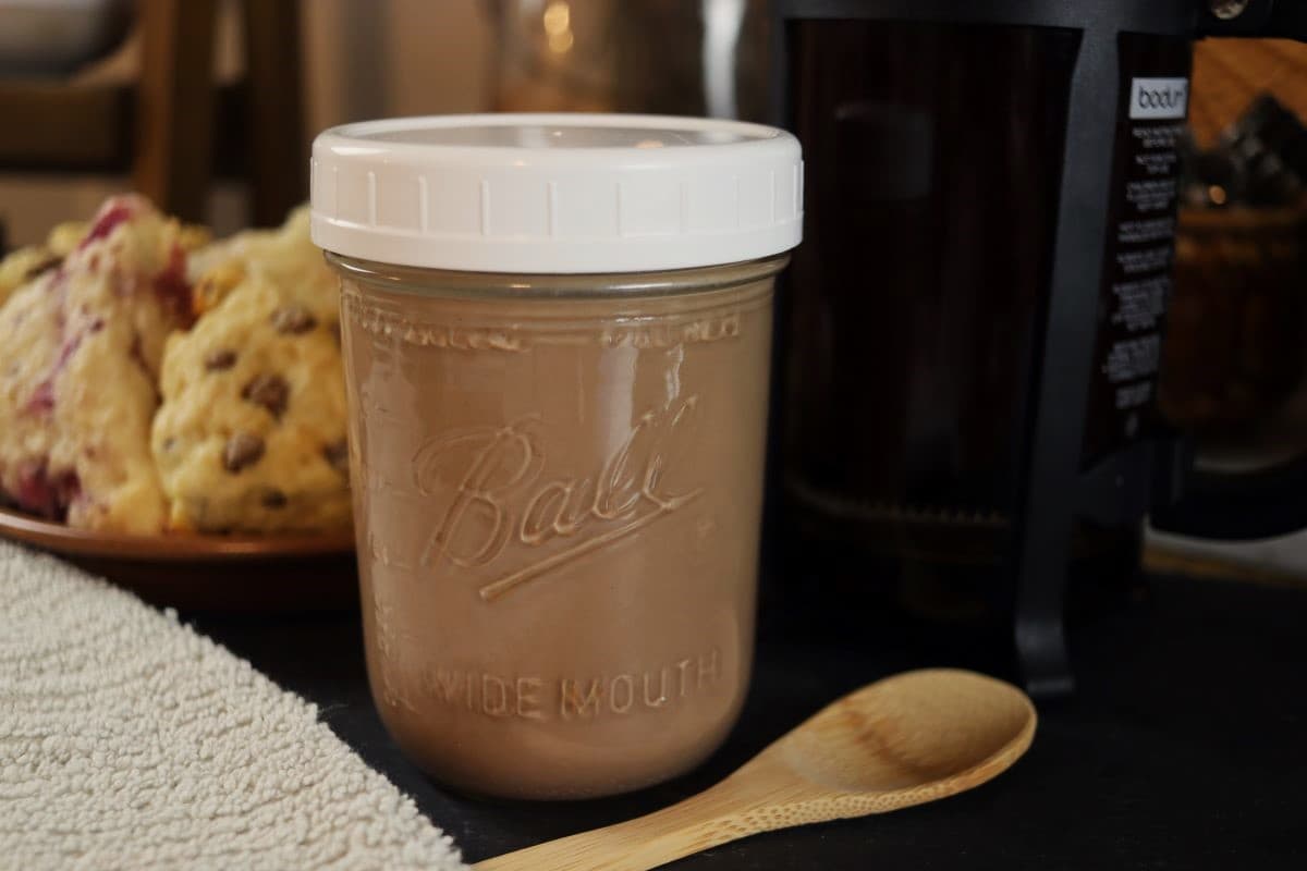 Pint size mason jar full of light brown liquid with a white lid on it sitting on a dark surface with chocolate chip scones visible in the background and a small wooden spoon in front.