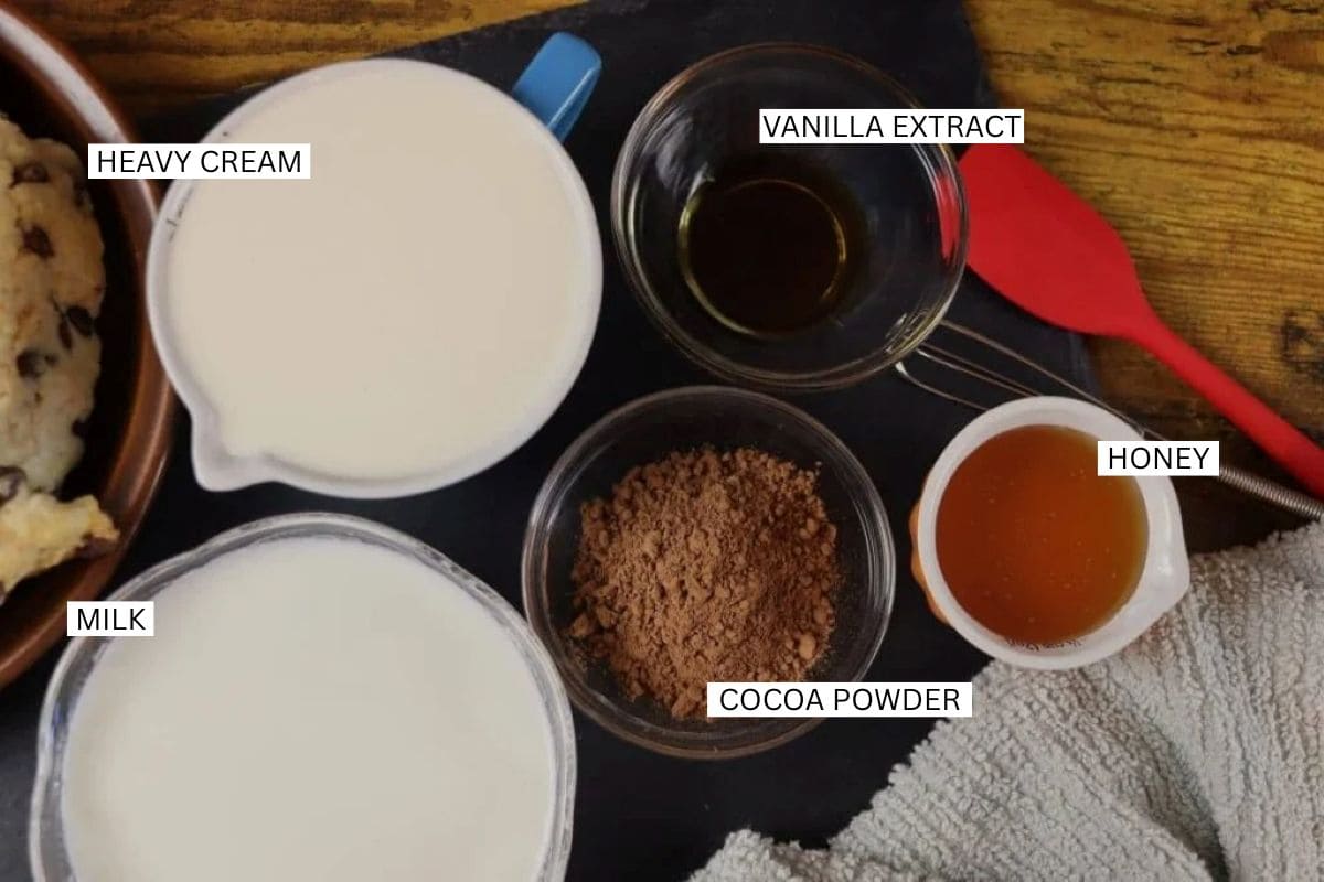 Labeled ingredients in bowls and cups: heavy cream, milk, cocoa powder, vanilla extract, and honey on a dark surface next to a red spatula.