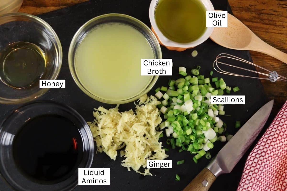 Top-down view of labeled ingredients on a dark cutting board: honey, chicken broth, olive oil, scallions, shredded ginger, liquid aminos, a wooden spoon, whisk, and a knife beside a red towel.