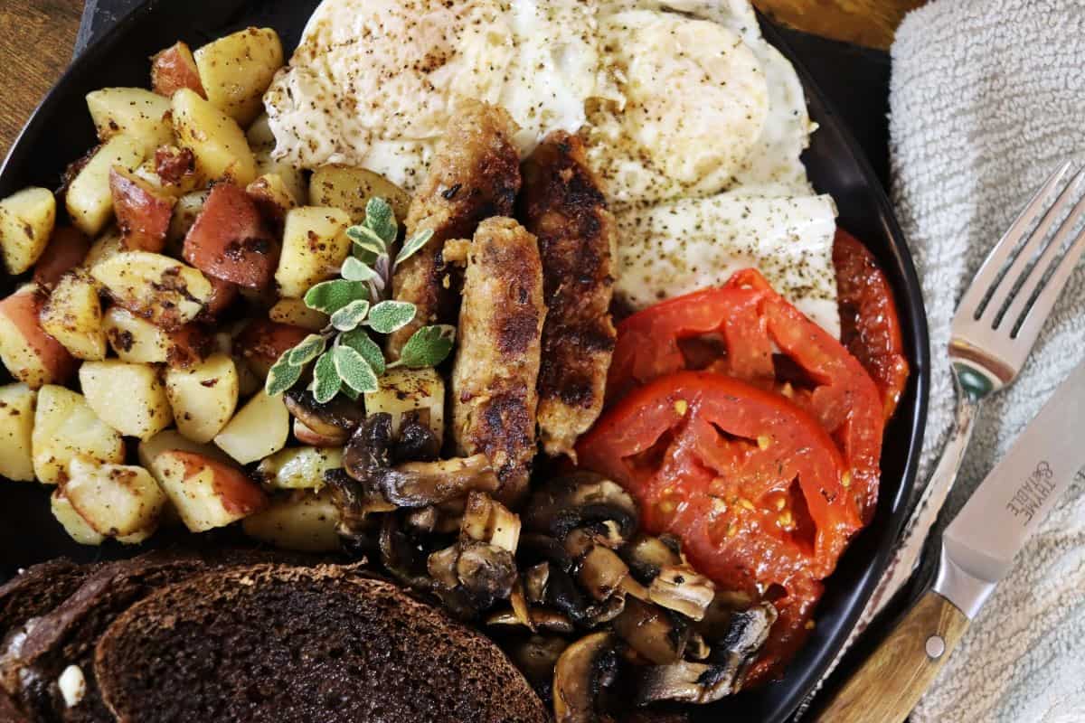 A breakfast plate with fried eggs, roasted potatoes, sausage, sliced tomatoes, cooked mushrooms, and dark rye bread. A fork and knife rest on a cloth napkin beside the plate.