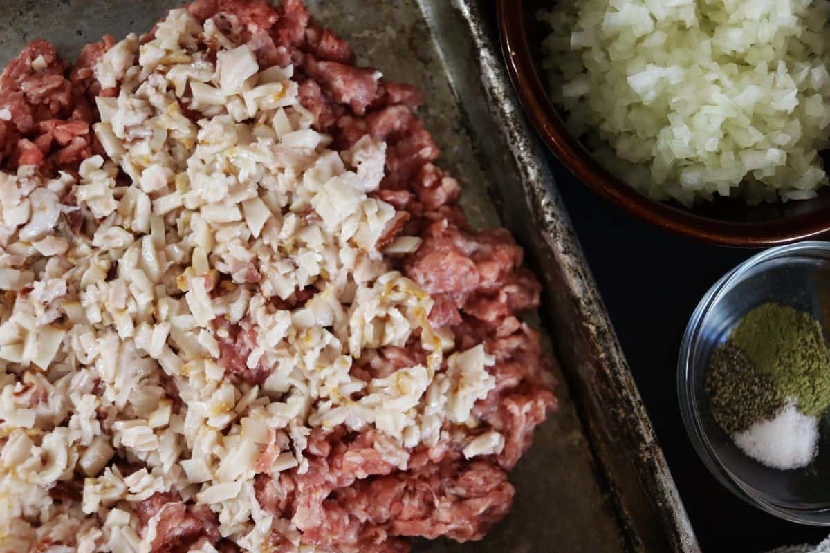 Raw ground pork spread on a sheet pan with chopped raw bacon fat on top of it and a dish of chopped onions visible to the side.
