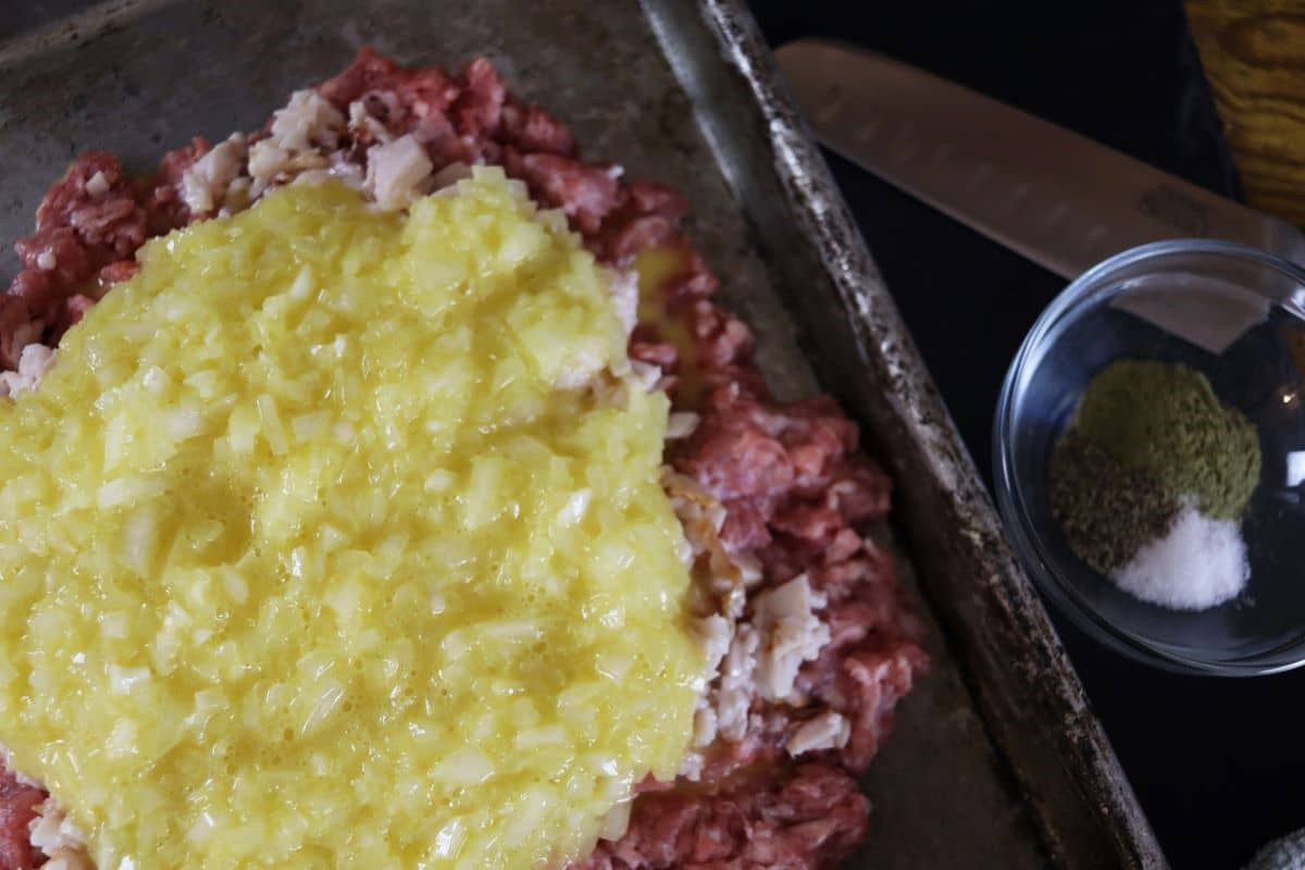 Raw ground meat spread on a baking sheet with beaten egg and onion mixture on top of it.