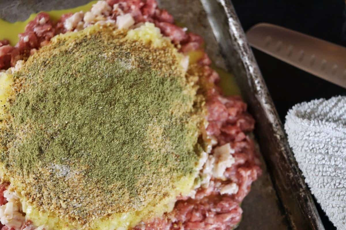 Raw ground meat spread on a sheet pan with a yellow mixture covered in seasonings spread on top of it.