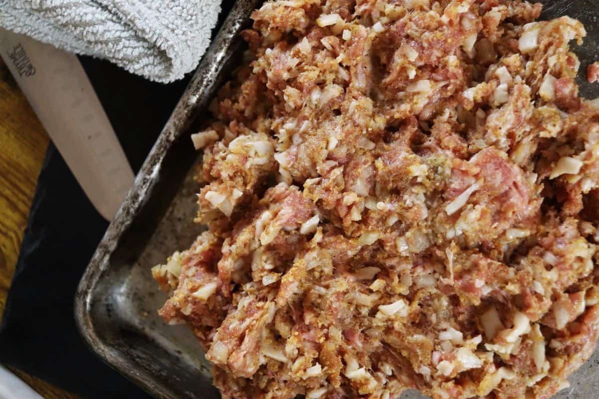 Raw ground meat mixture with seasoning and diced onions visible in it spread on a sheet pan.