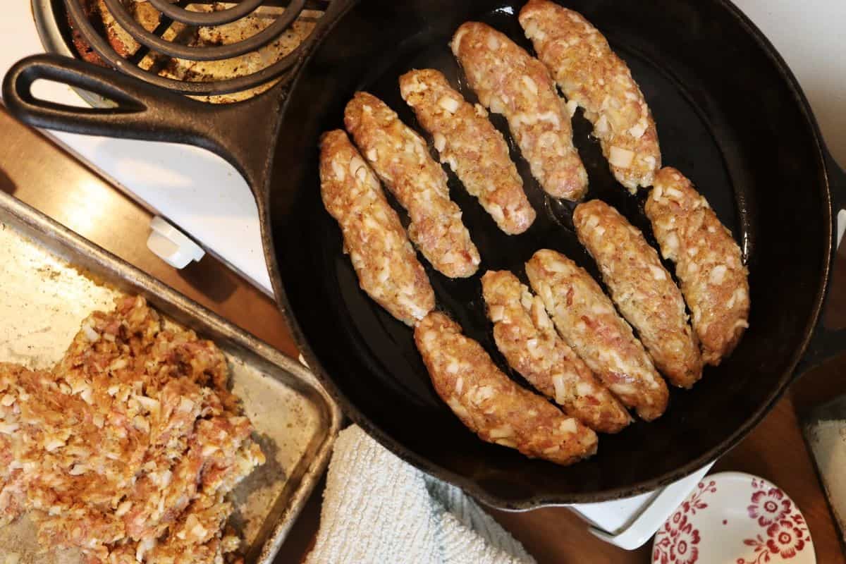 Raw sausage links in a cast iron skillet on a stovetop with the edge of a sheet pan with raw meat mixture on it visible to the side.