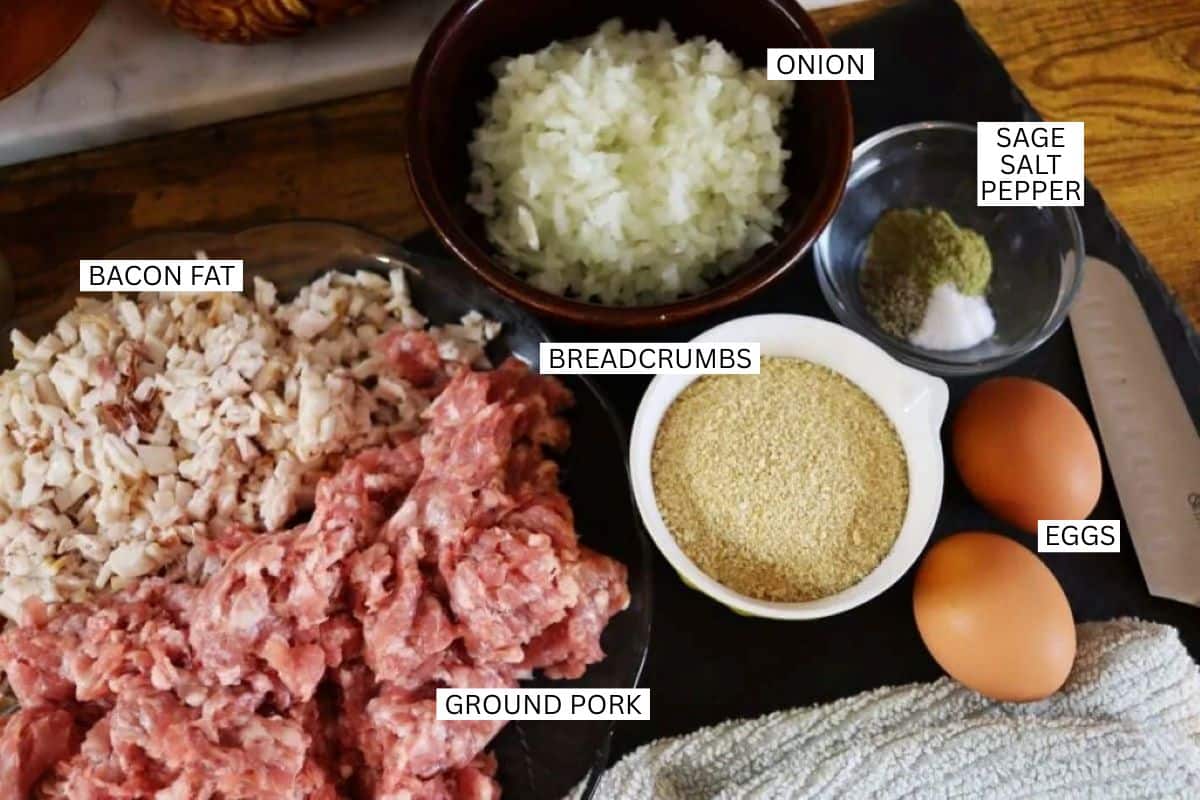 Chopped onion, sage, salt, pepper, breadcrumbs, eggs, chopped bacon fat, and ground pork are arranged separately on a table, each labeled with their ingredient name.