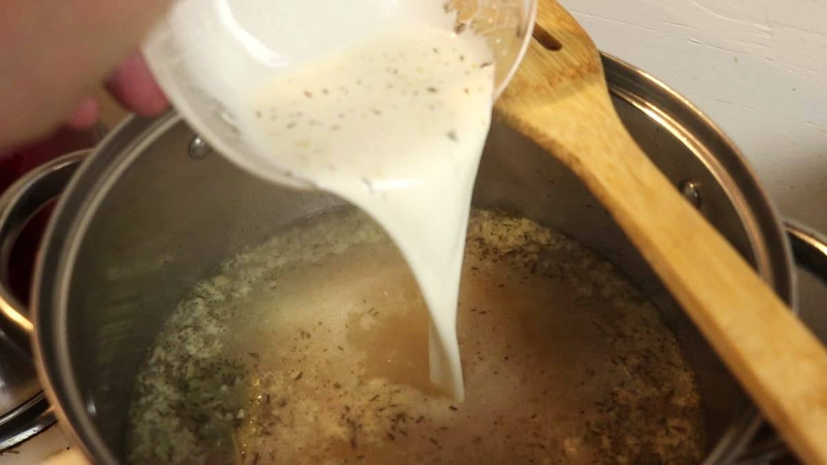 White sauce with seasoning in it pouring into a pot of golden broth.