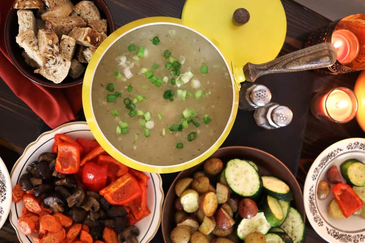 Fondue of golden broth topped with scallions sitting on a table surrounded by bowls of bread and vegetables, a salt and pepper shaker, and the lid for the pot.