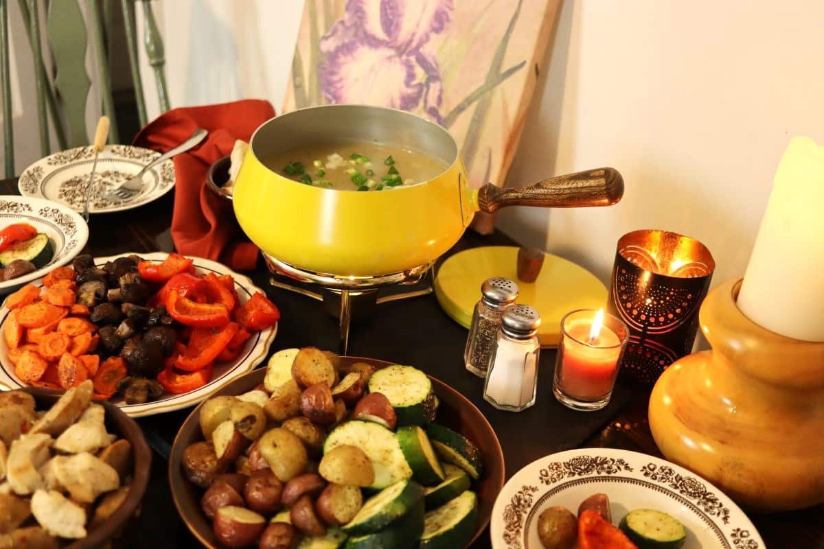 A table set with a yellow fondue pot filled with broth, surrounded by plates of assorted vegetables, bread cubes, and dipping items. A lit candle, salt and pepper shakers, and a painted flower are in the background.