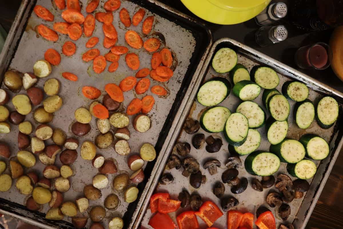 Chunks of carrots, potatoes, zucchini, mushrooms, and red bell pepper seasoned and spread out on baking sheets.
